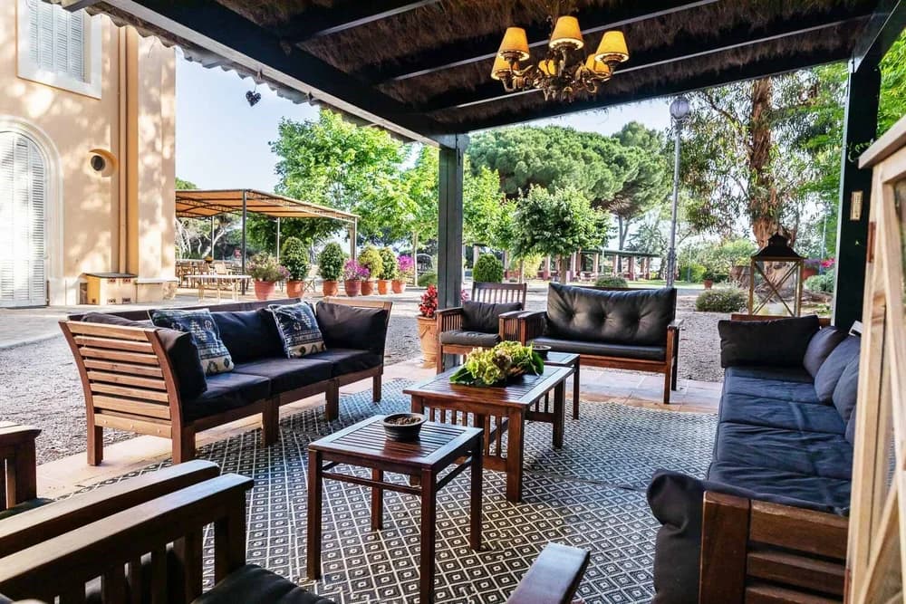 An outdoor lounge under a reed-roofed veranda, with dark cushioned sofas facing the garden