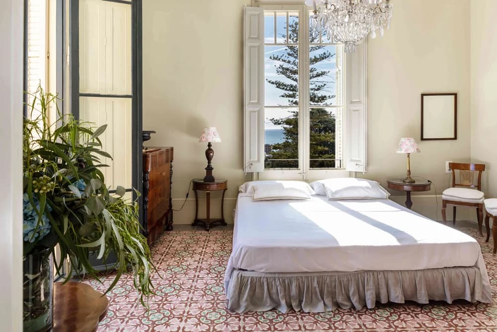 A bedroom with patterned tile floors, a crystal chandelier and shuttered French doors opening to a sea view