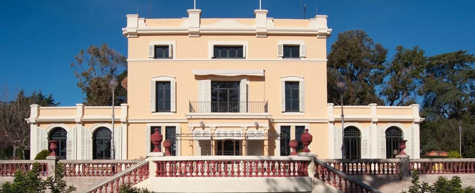 The front facade of Villa Blanca, a cream-colored villa with a red stone balustrade, under a clear blue sky